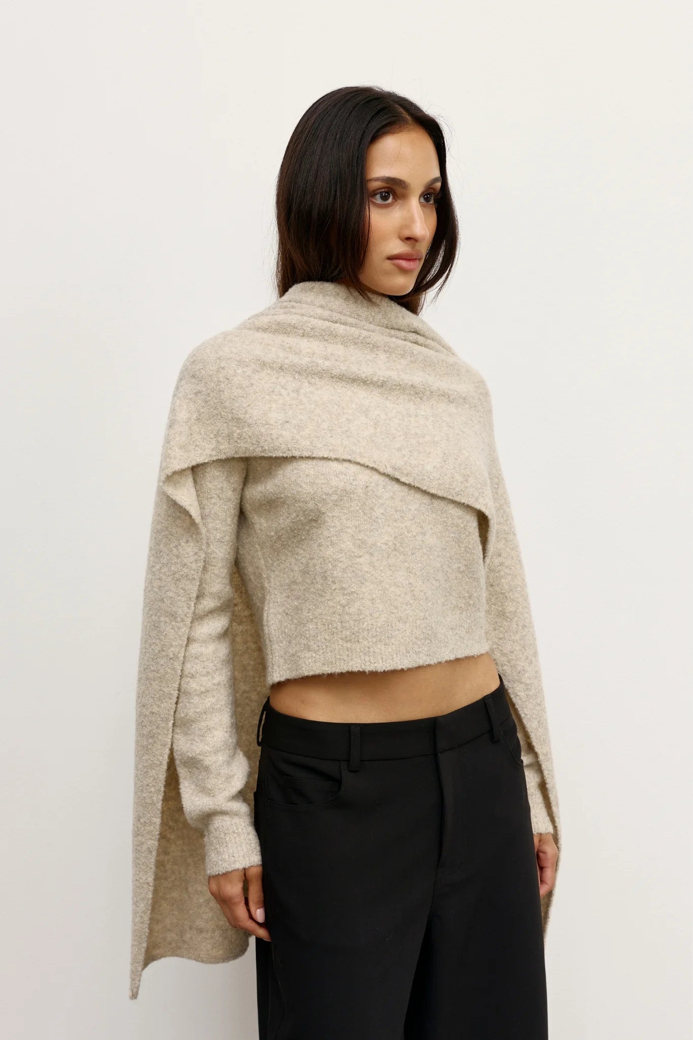 Woman wearing a beige knitted shawl over a cropped top and black pants on a white background