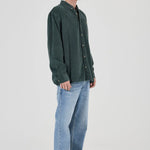 A man wearing a dark green corduroy button-up shirt, light blue jeans, and gray sneakers, standing in profile against a light gray studio background.