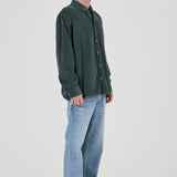A man wearing a dark green corduroy button-up shirt, light blue jeans, and gray sneakers, standing in profile against a light gray studio background.