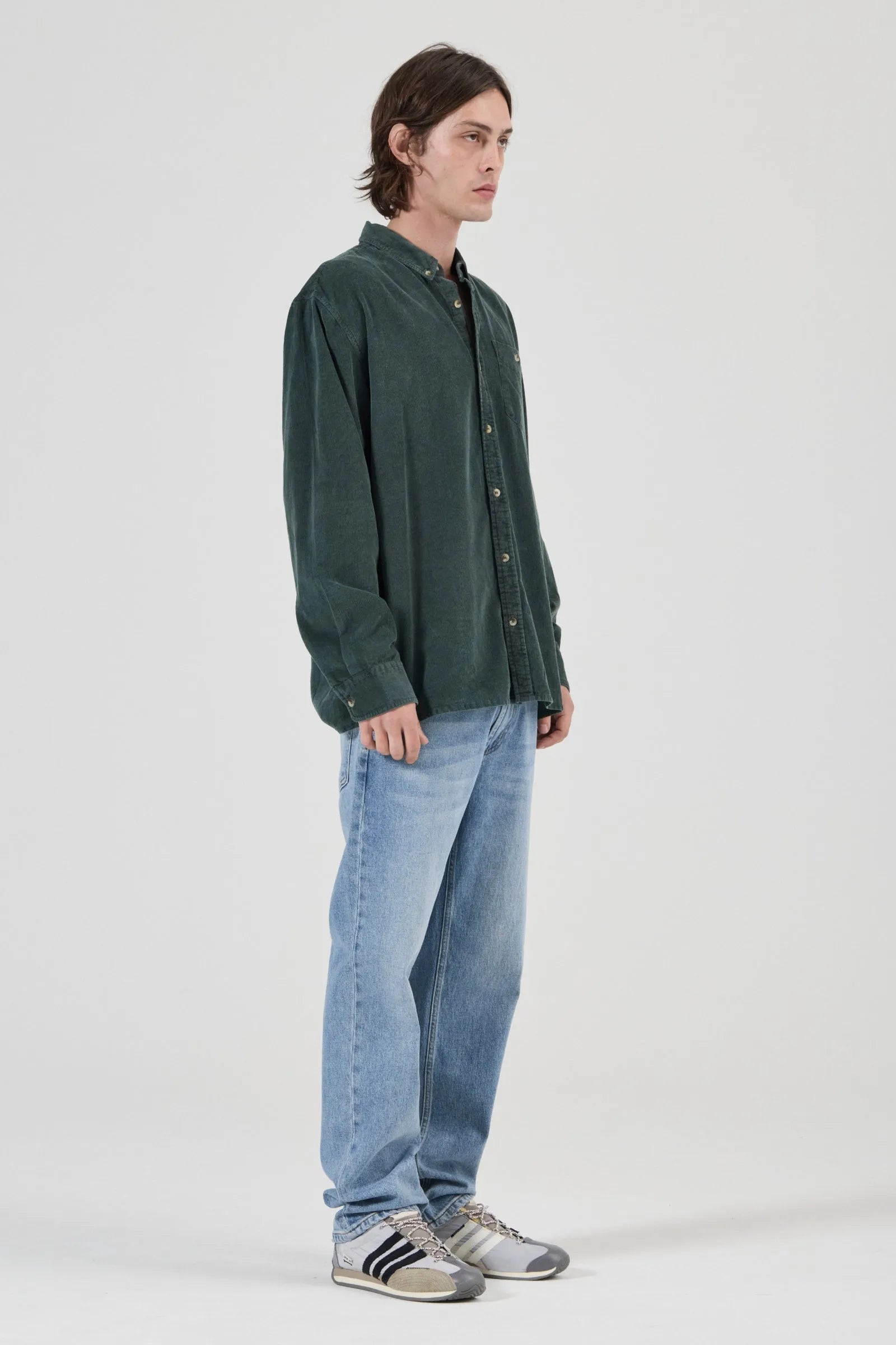 A man wearing a dark green corduroy button-up shirt, light blue jeans, and gray sneakers, standing in profile against a light gray studio background.