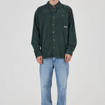 A man modeling a dark green corduroy button-up shirt with light blue jeans and gray sneakers, standing against a light gray studio background.
