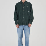 A man modeling a dark green corduroy button-up shirt with light blue jeans and gray sneakers, standing against a light gray studio background.