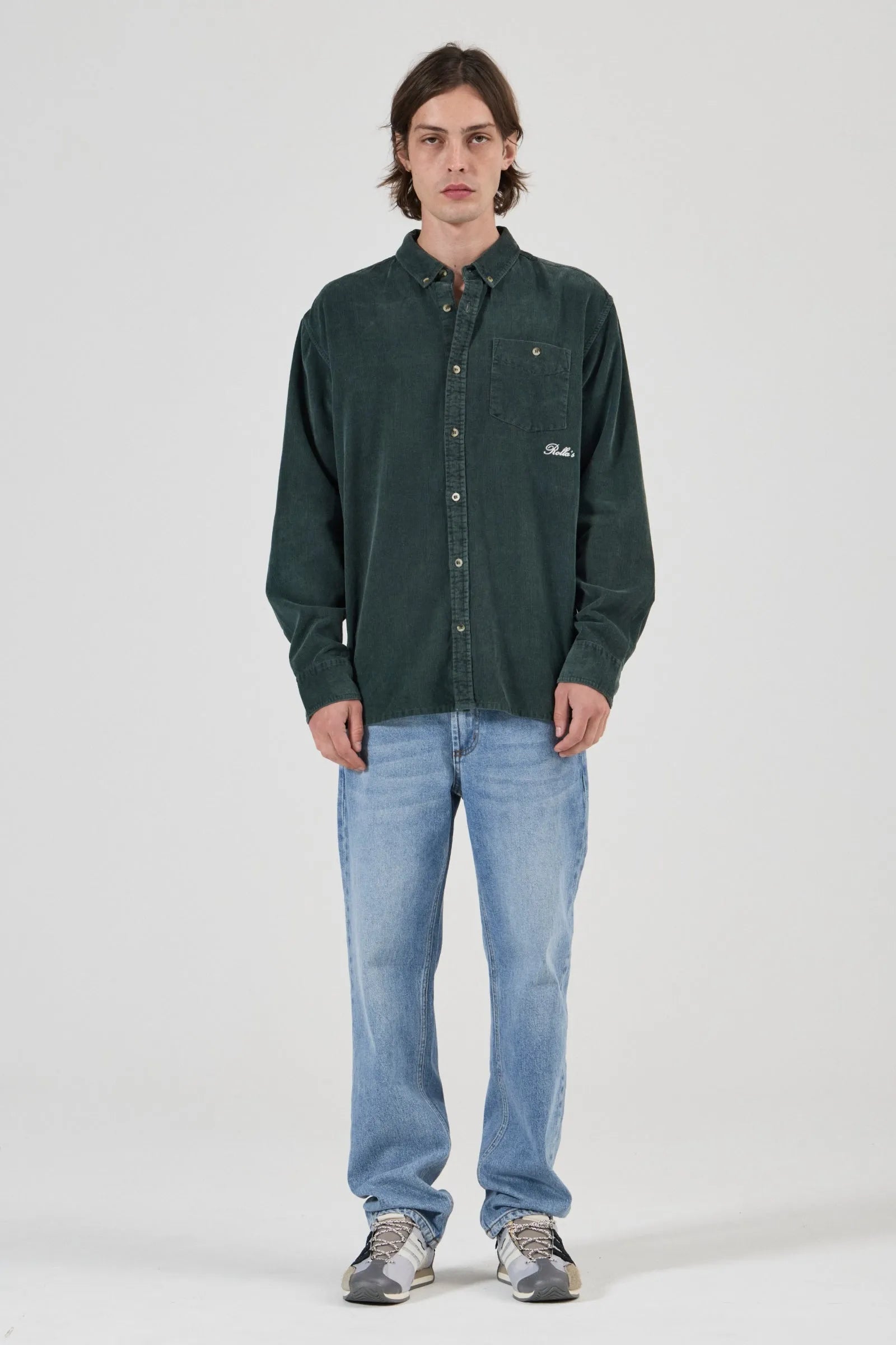 A man modeling a dark green corduroy button-up shirt with light blue jeans and gray sneakers, standing against a light gray studio background.
