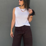 A woman wearing a white ribbed sleeveless top and dark brown wide-leg pants, standing against a textured gray wall.
