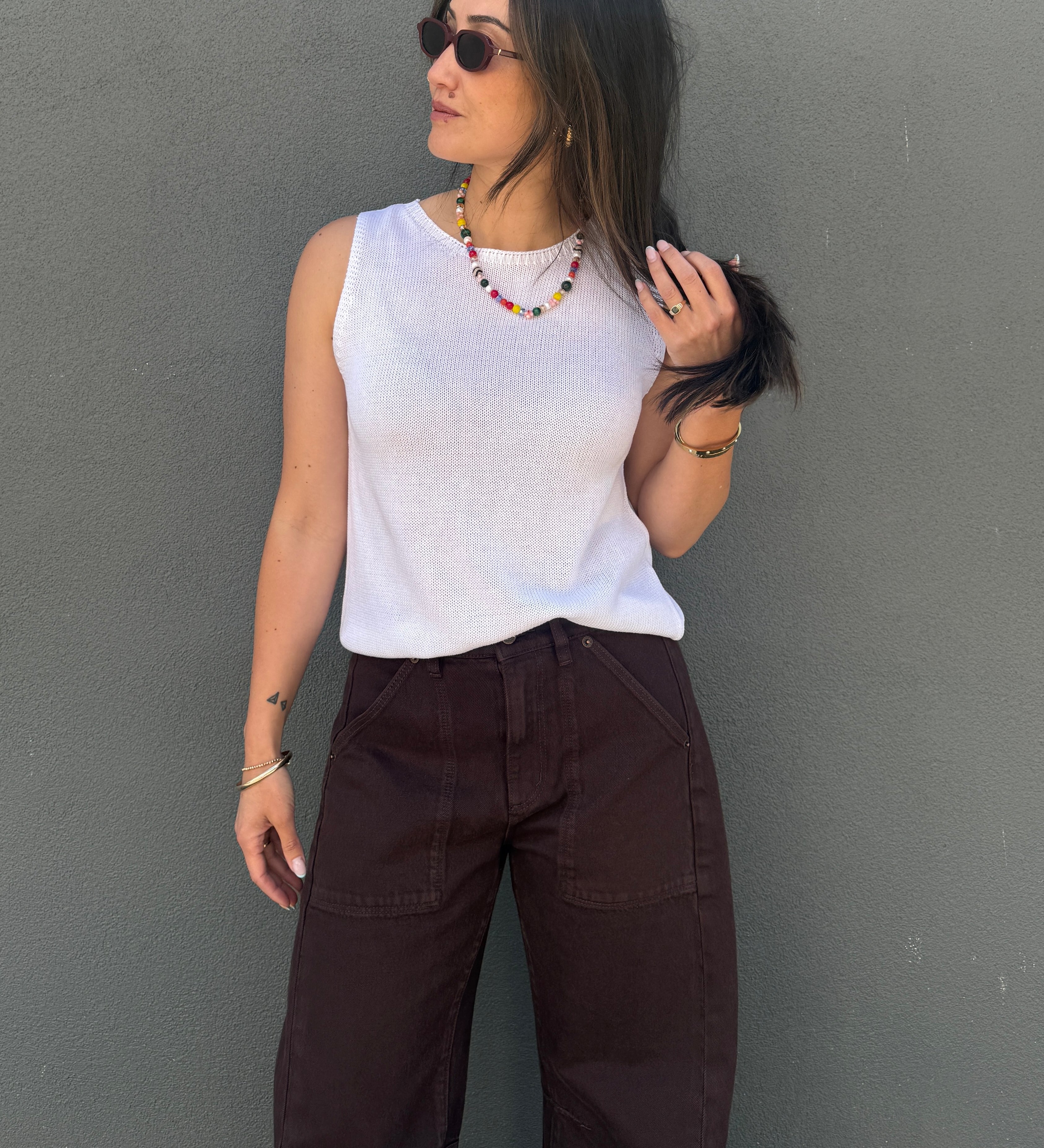 A woman wearing a white ribbed sleeveless top and dark brown wide-leg pants, standing against a textured gray wall.