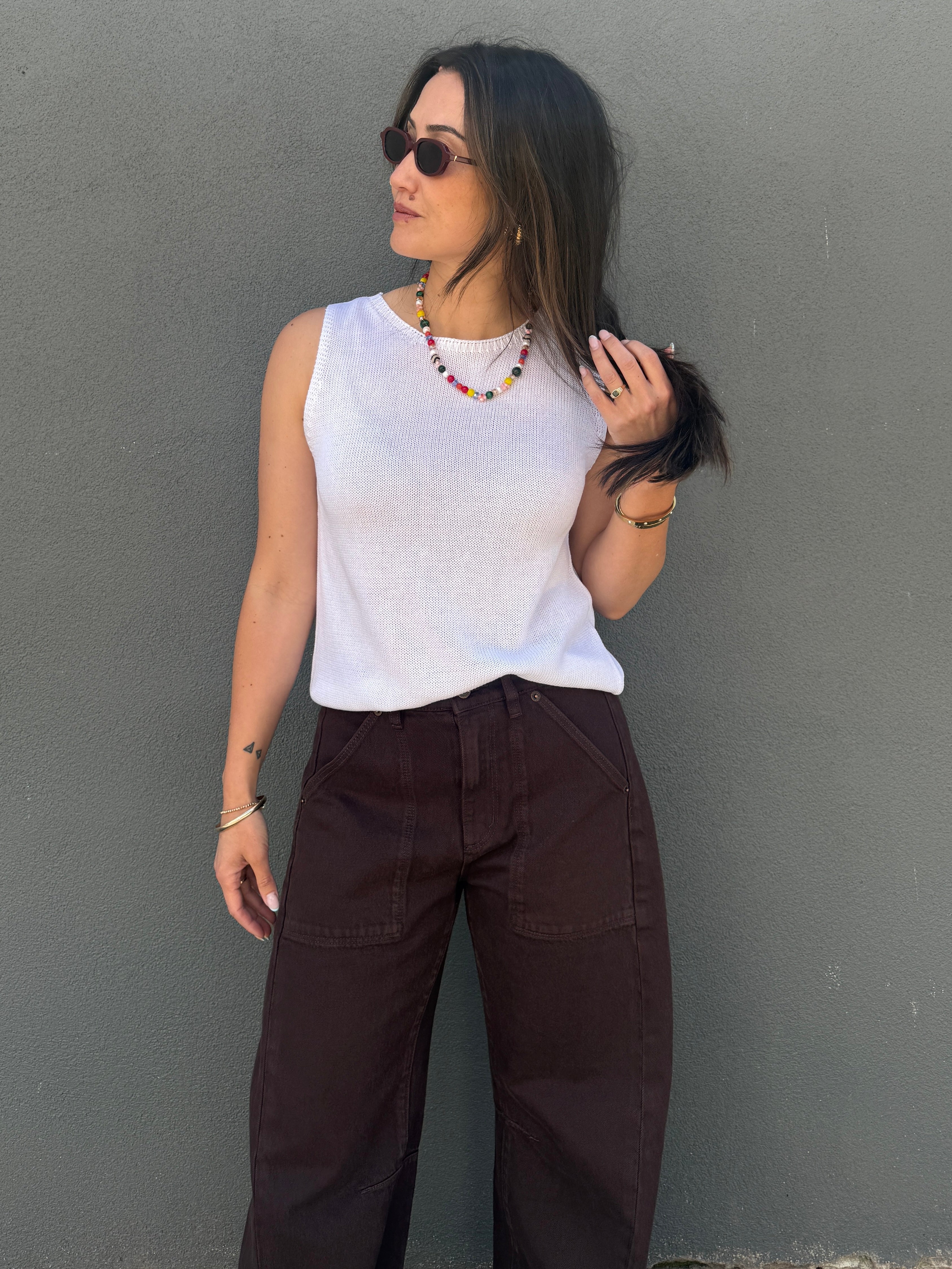 A woman wearing a white ribbed sleeveless top and dark brown wide-leg pants, standing against a textured gray wall.