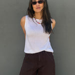 A woman wearing a white ribbed sleeveless top and dark brown wide-leg pants, standing against a textured gray wall.