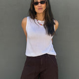 A woman wearing a white ribbed sleeveless top and dark brown wide-leg pants, standing against a textured gray wall.