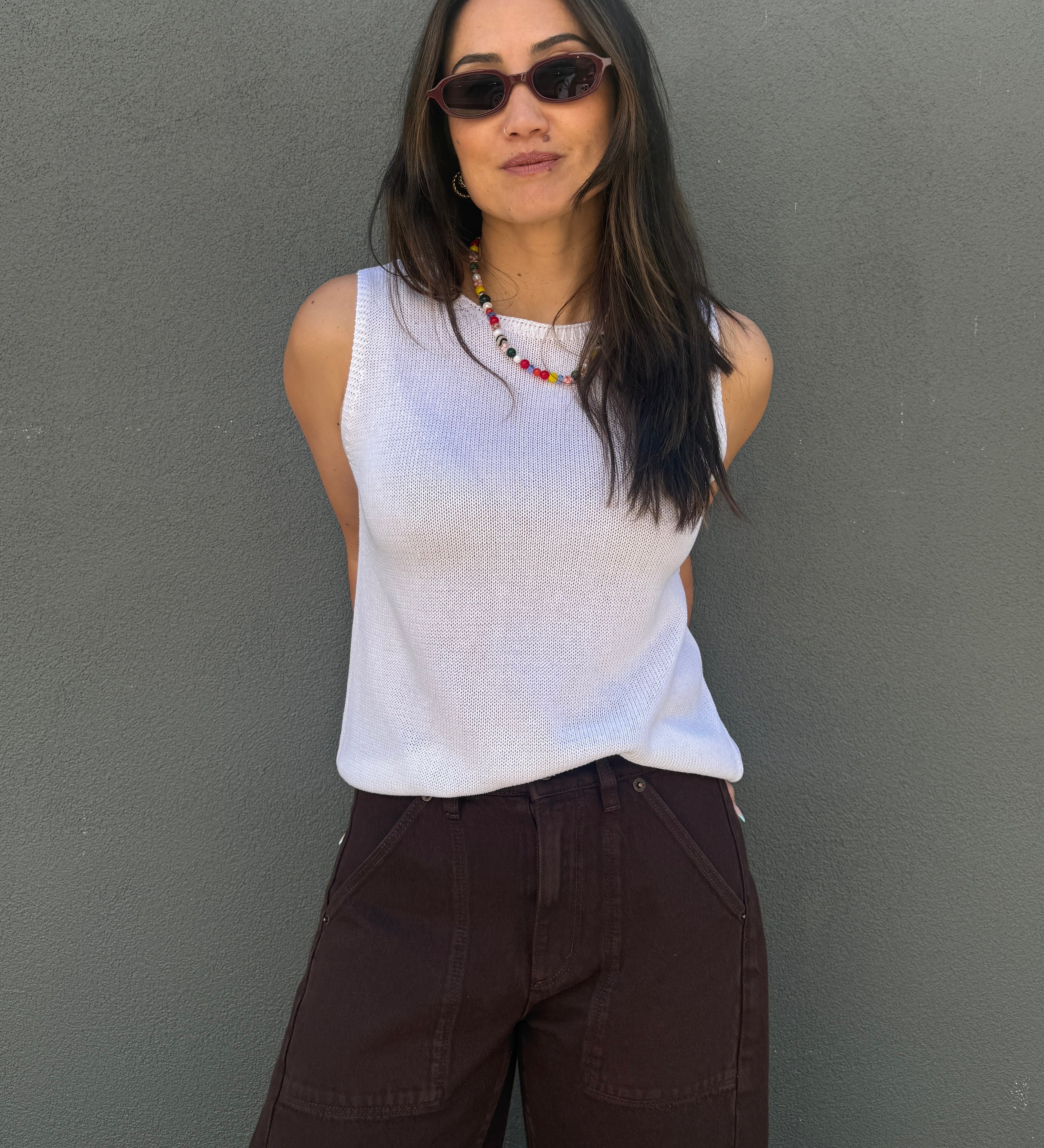 A woman wearing a white ribbed sleeveless top and dark brown wide-leg pants, standing against a textured gray wall.