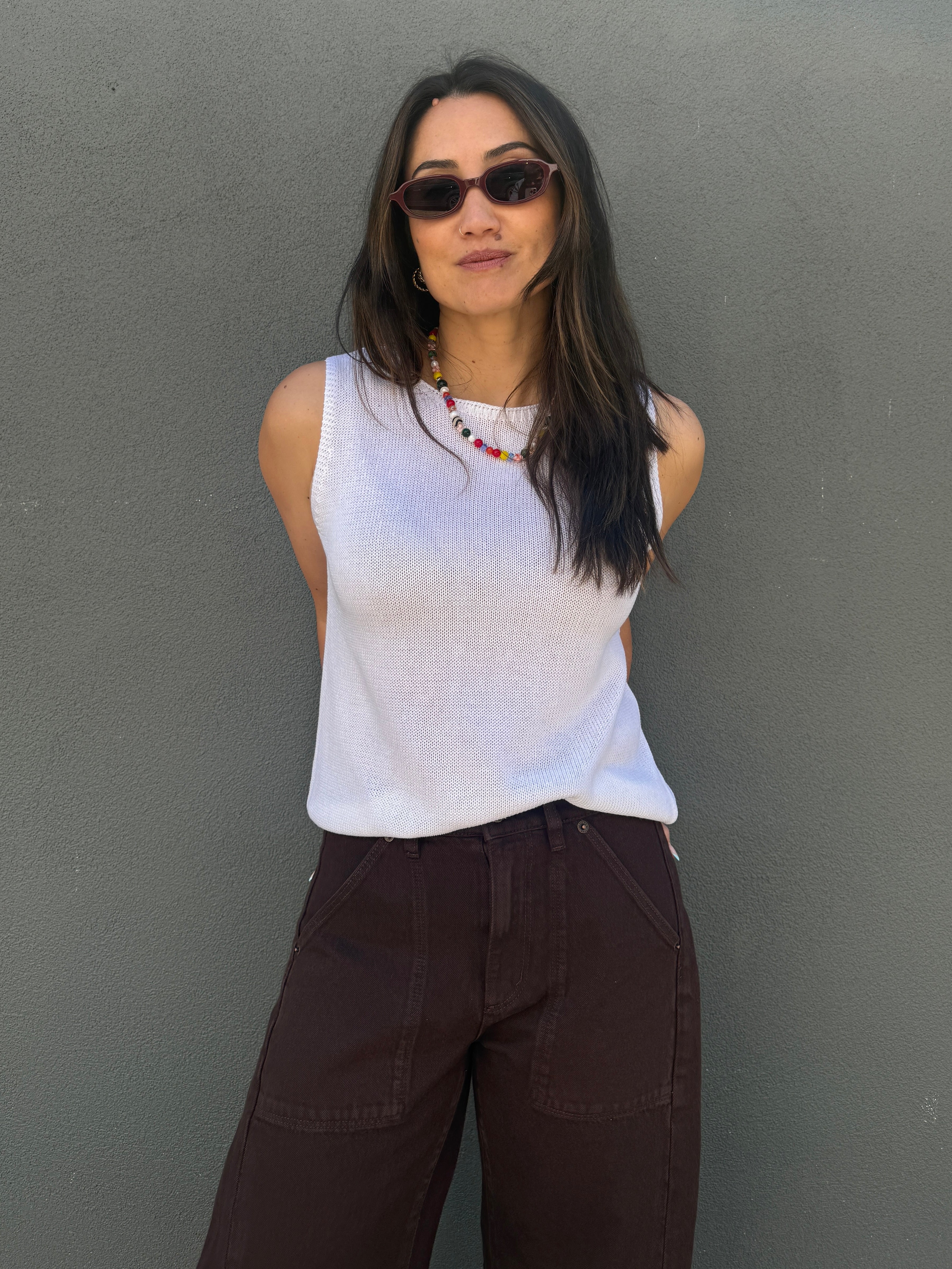 A woman wearing a white ribbed sleeveless top and dark brown wide-leg pants, standing against a textured gray wall.