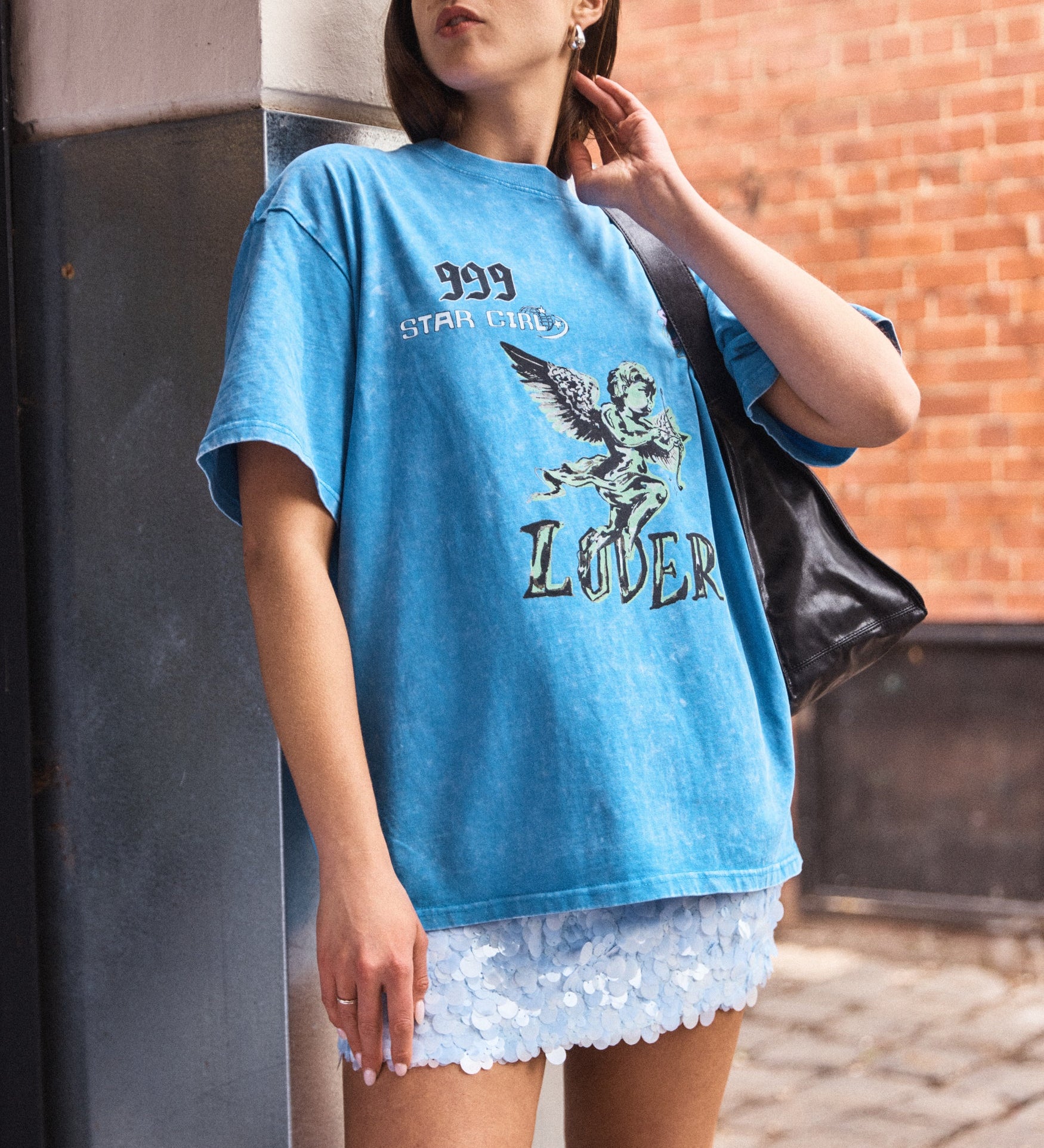 Woman wearing a blue graphic t-shirt and white shorts standing against a brick wall.