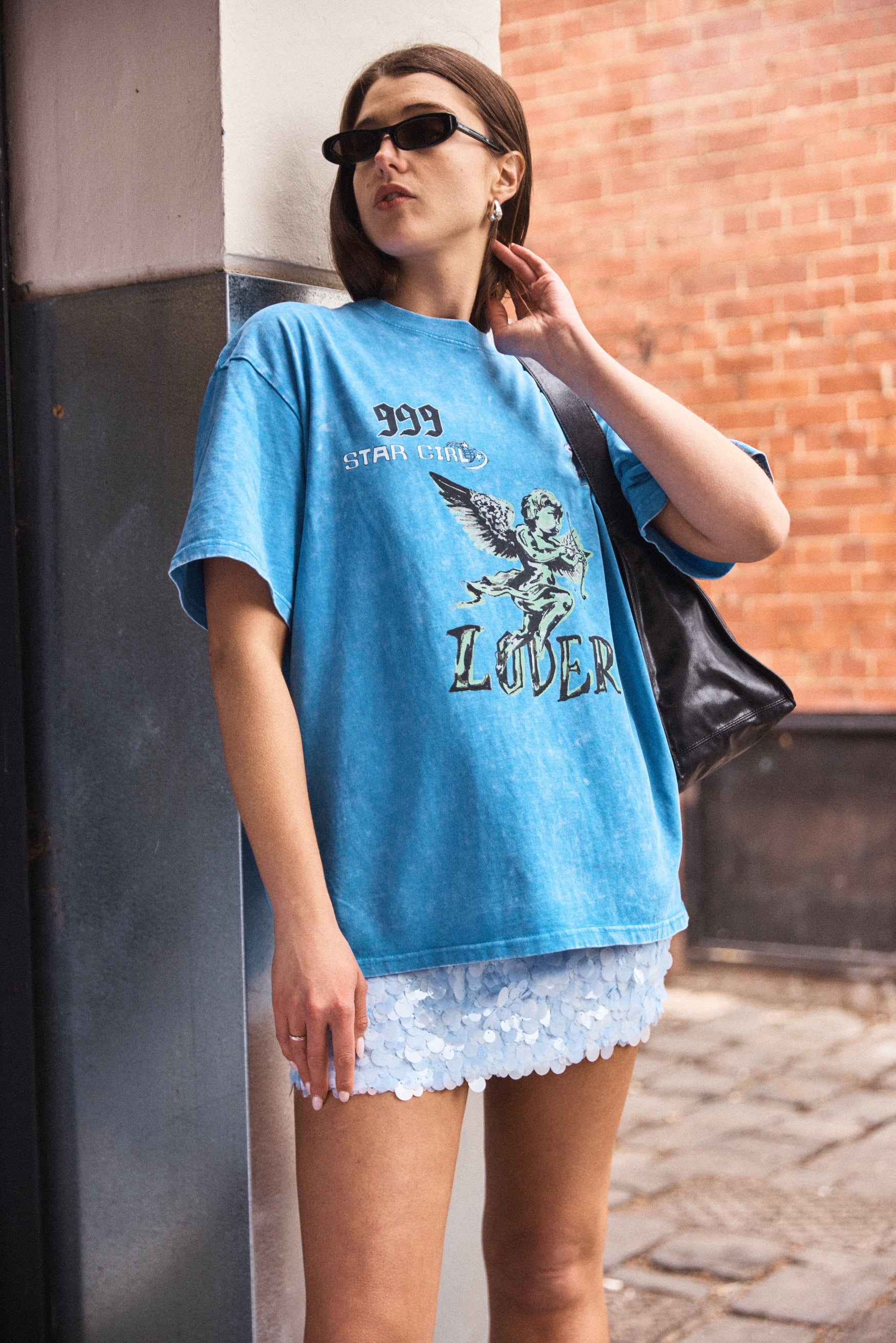 Woman wearing a blue graphic t-shirt and white shorts standing against a brick wall.