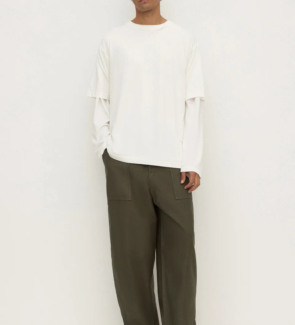 A man modeling a white long-sleeve top with layered ruffle details on the sleeves and wide-leg olive green trousers, standing against a plain white studio background.