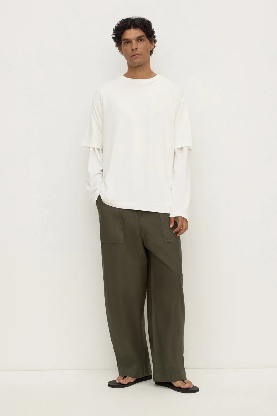 A man modeling a white long-sleeve top with layered ruffle details on the sleeves and wide-leg olive green trousers, standing against a plain white studio background.