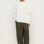 A man modeling a white long-sleeve top with layered ruffle details on the sleeves and wide-leg olive green trousers, standing against a plain white studio background.