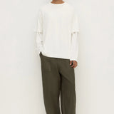 A man modeling a white long-sleeve top with layered ruffle details on the sleeves and wide-leg olive green trousers, standing against a plain white studio background.