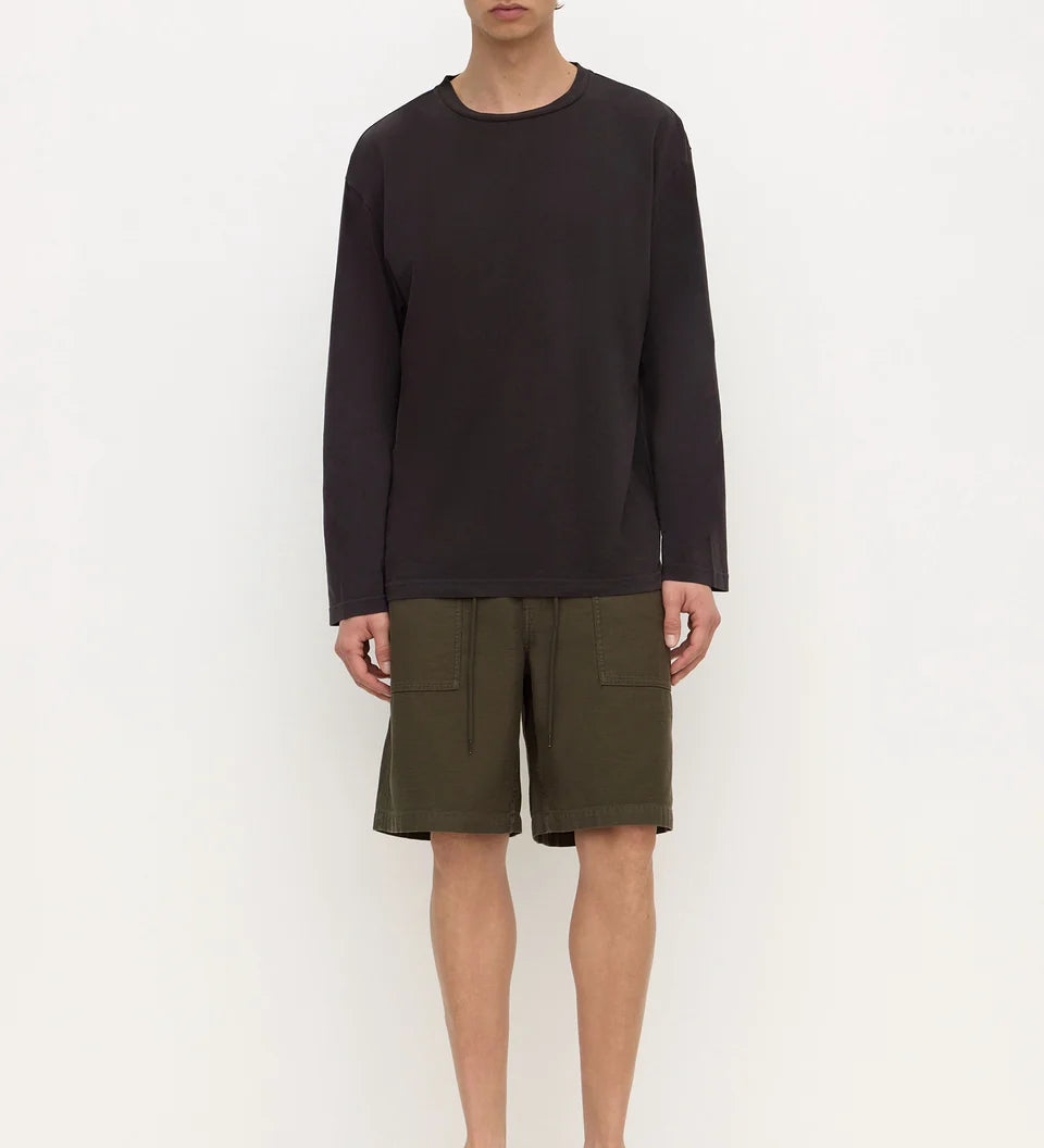 A man stands against a plain white studio background wearing a dark charcoal long-sleeve crewneck shirt, olive green shorts, and black slip-on shoes.