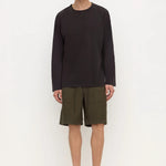 A man stands against a plain white studio background wearing a dark charcoal long-sleeve crewneck shirt, olive green shorts, and black slip-on shoes.