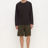 A man stands against a plain white studio background wearing a dark charcoal long-sleeve crewneck shirt, olive green shorts, and black slip-on shoes.