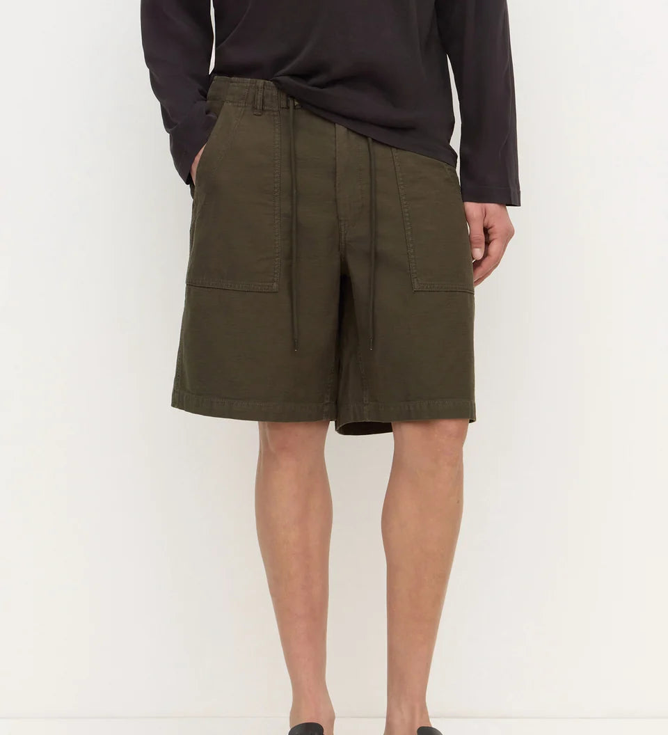 A model wearing olive green drawstring cargo shorts with side pockets, paired with a dark long-sleeve shirt and black clogs, standing against a plain white studio background.