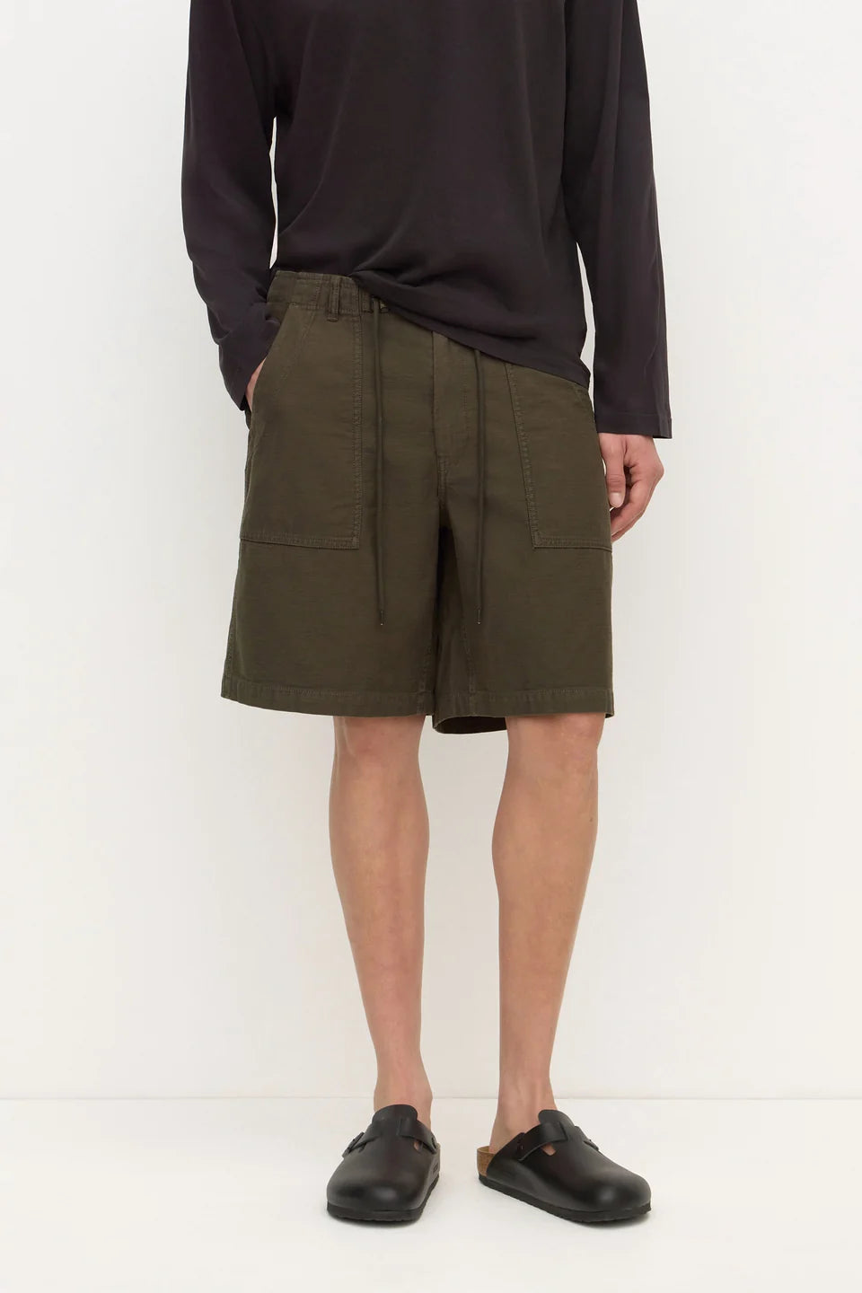 A model wearing olive green drawstring cargo shorts with side pockets, paired with a dark long-sleeve shirt and black clogs, standing against a plain white studio background.