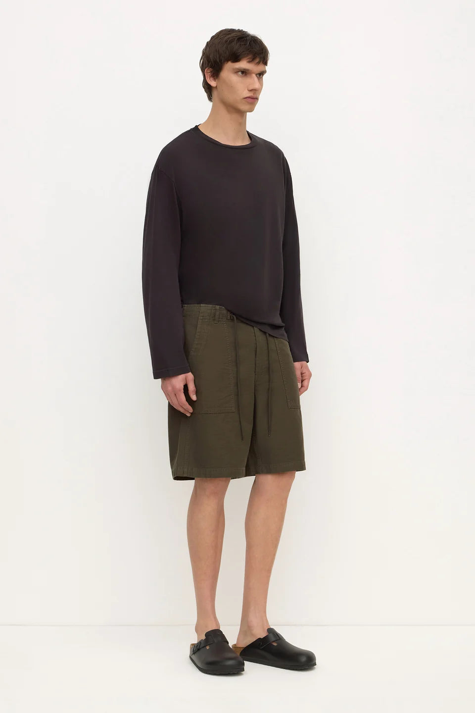 A model stands against a plain white studio background wearing a dark charcoal long-sleeve top, olive green shorts, and black clogs.