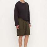 A model stands against a plain white studio background wearing a dark charcoal long-sleeve top, olive green shorts, and black clogs.