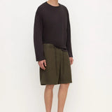 A model stands against a plain white studio background wearing a dark charcoal long-sleeve top, olive green shorts, and black clogs.