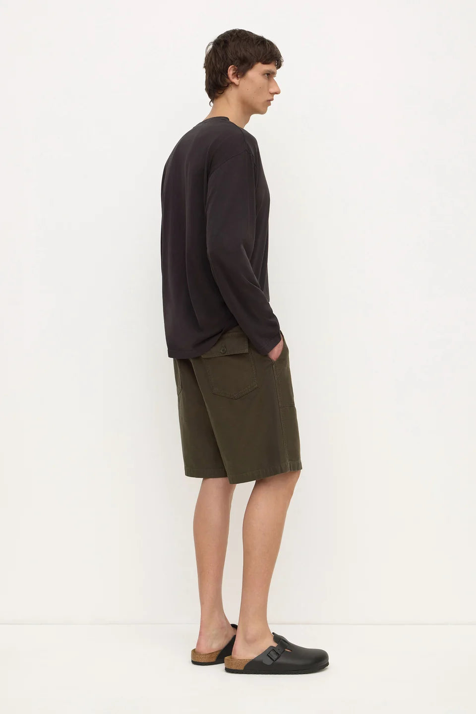 A man stands in profile wearing a dark charcoal long-sleeve shirt, olive green shorts, and black clogs against a plain white studio background.