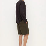 A man stands in profile wearing a dark charcoal long-sleeve shirt, olive green shorts, and black clogs against a plain white studio background.