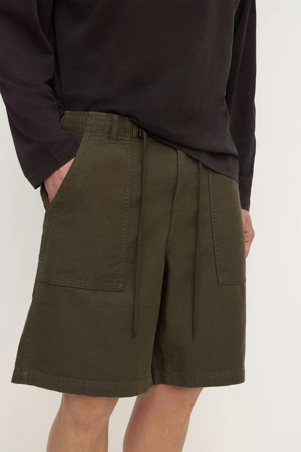 A person wearing olive green drawstring shorts with large front patch pockets and a dark long-sleeve top, photographed against a plain light background.