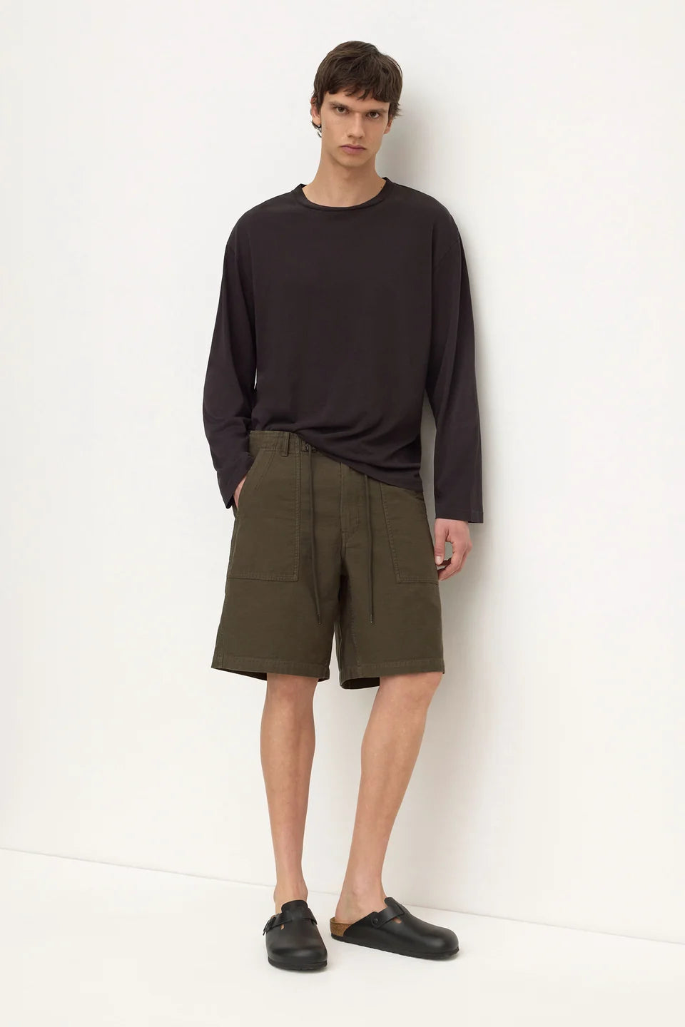 A man stands against a plain white wall wearing a dark charcoal long-sleeve shirt, olive green utility shorts, and black slip-on shoes.