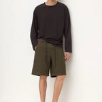 A man stands against a plain white wall wearing a dark charcoal long-sleeve shirt, olive green utility shorts, and black slip-on shoes.