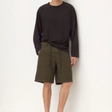 A man stands against a plain white wall wearing a dark charcoal long-sleeve shirt, olive green utility shorts, and black slip-on shoes.