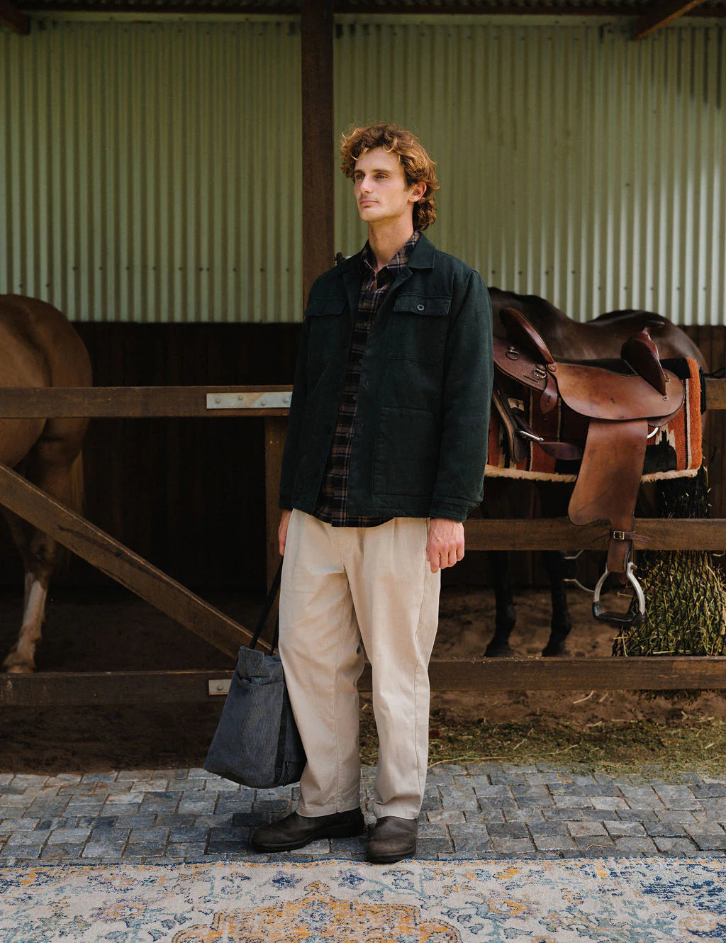 Man standing in a stable with horses and saddles in the background