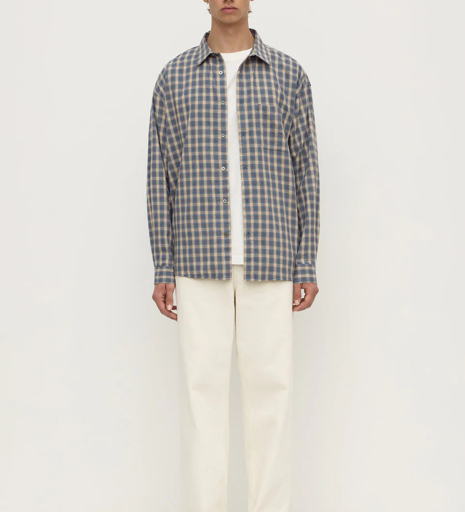 A man stands against a plain white studio background wearing a gray and white plaid button-down shirt over a white t-shirt, cream-colored trousers, and white sneakers.