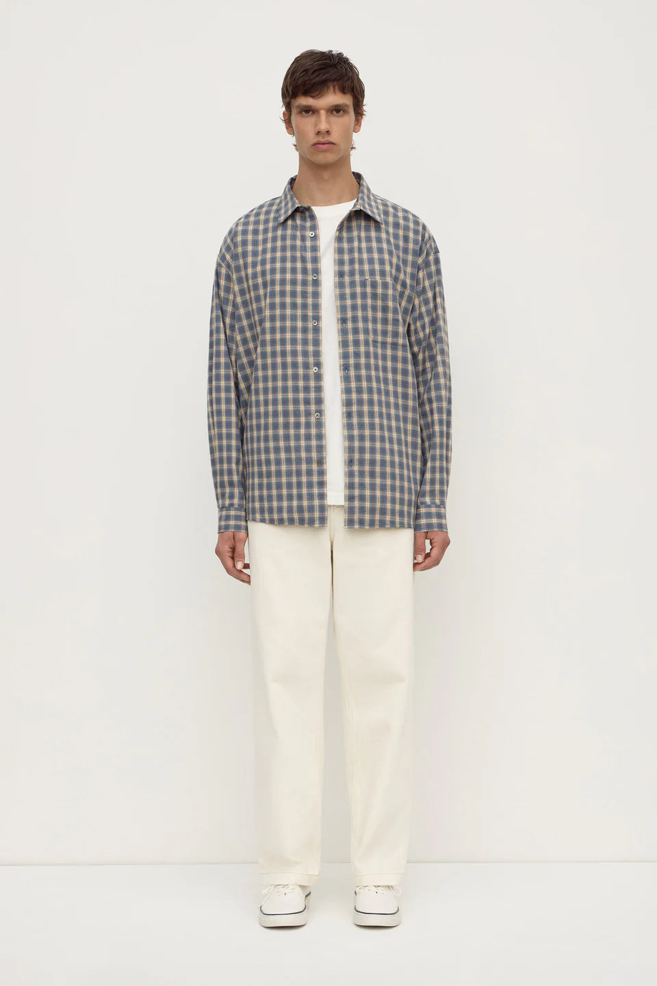 A man stands against a plain white studio background wearing a gray and white plaid button-down shirt over a white t-shirt, cream-colored trousers, and white sneakers.