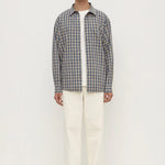 A man stands against a plain white studio background wearing a gray and white plaid button-down shirt over a white t-shirt, cream-colored trousers, and white sneakers.