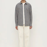 A man stands against a plain white studio background wearing a gray and white plaid button-down shirt over a white t-shirt, cream-colored trousers, and white sneakers.
