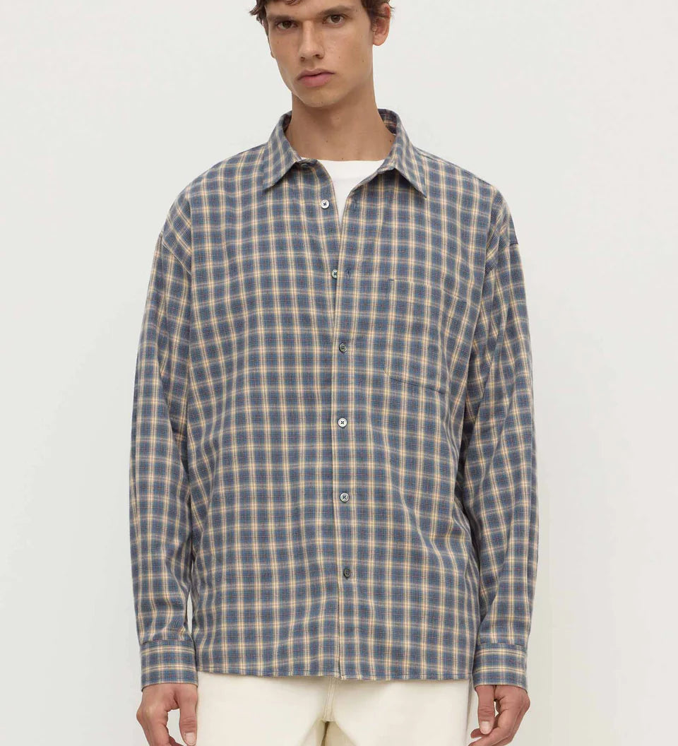 A man wearing a long-sleeve button-up plaid shirt in blue and beige over a white undershirt, paired with cream trousers, against a plain off-white studio background.