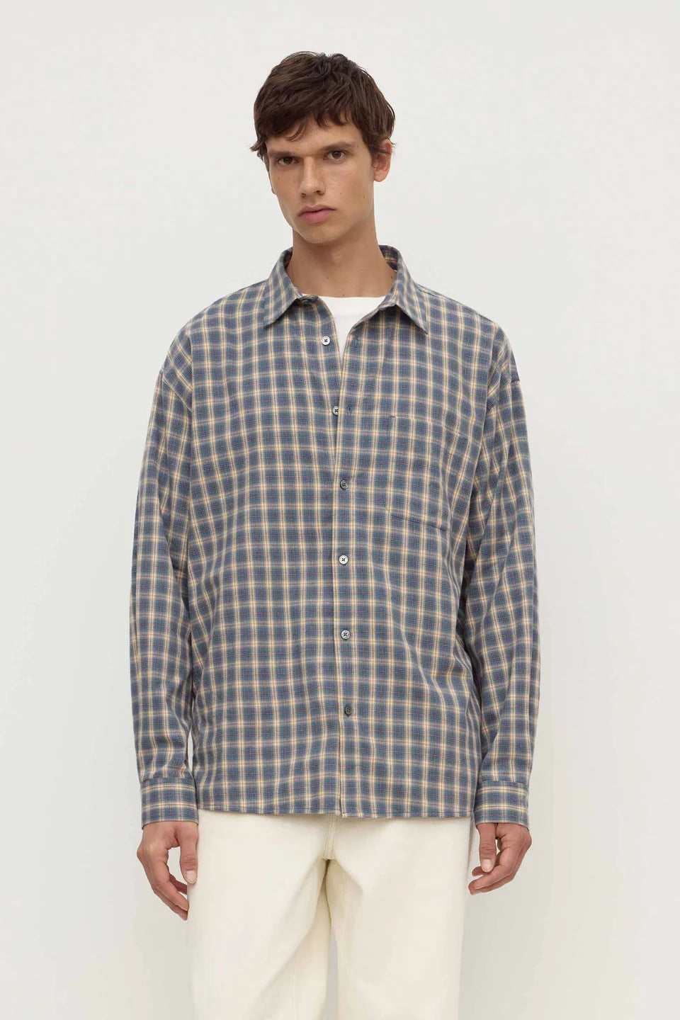 A man wearing a long-sleeve button-up plaid shirt in blue and beige over a white undershirt, paired with cream trousers, against a plain off-white studio background.