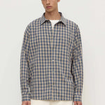 A man wearing a long-sleeve button-up plaid shirt in blue and beige over a white undershirt, paired with cream trousers, against a plain off-white studio background.