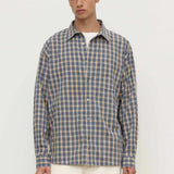 A man wearing a long-sleeve button-up plaid shirt in blue and beige over a white undershirt, paired with cream trousers, against a plain off-white studio background.