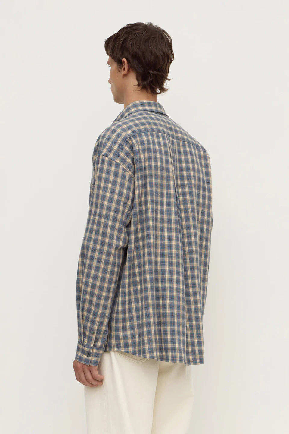 A man shown from the back wearing a blue and beige plaid button-up shirt and cream trousers, photographed against a plain white studio background.