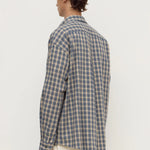 A man shown from the back wearing a blue and beige plaid button-up shirt and cream trousers, photographed against a plain white studio background.