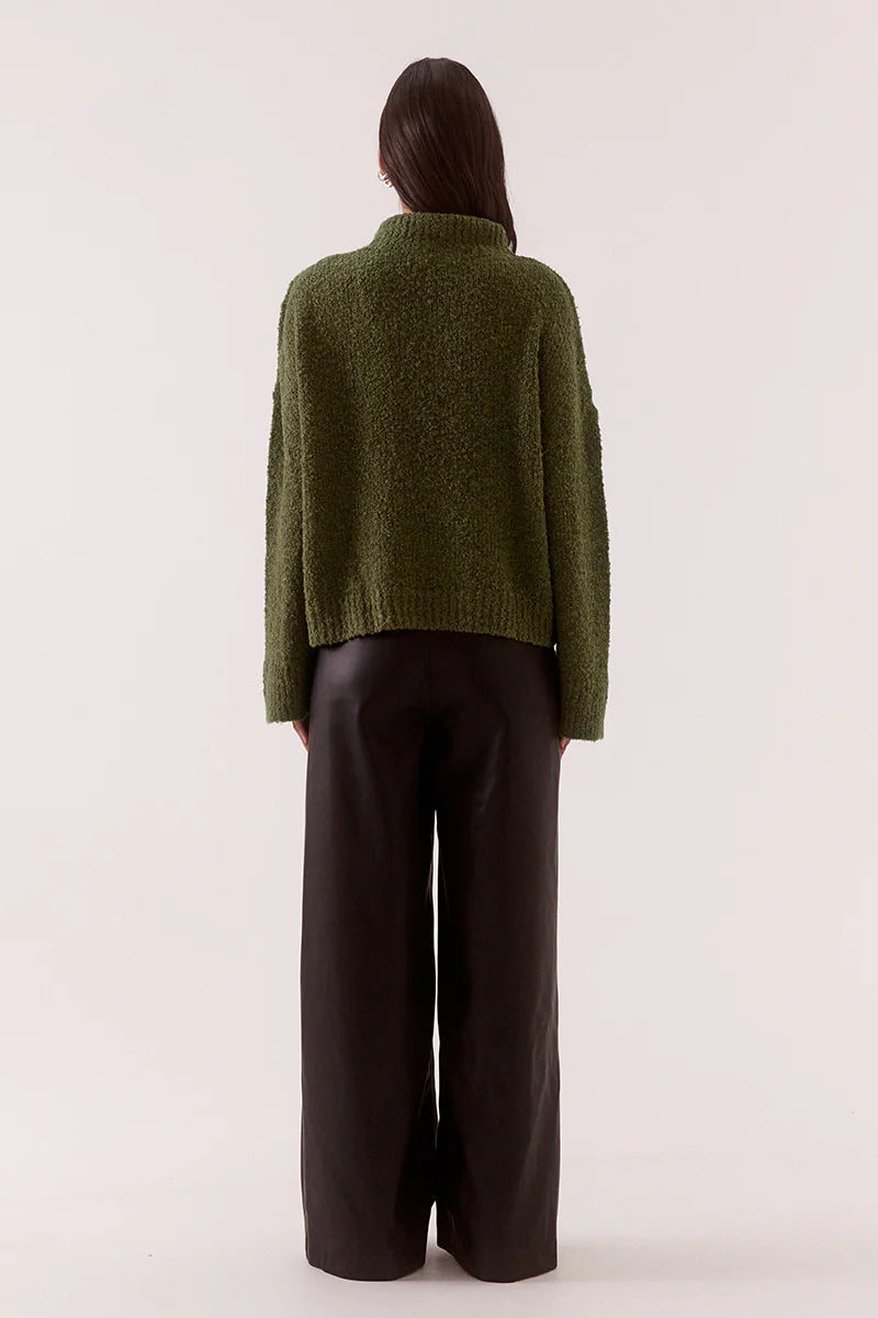 Person wearing a green sweater and dark pants on a white background