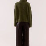 Person wearing a green sweater and dark pants on a white background
