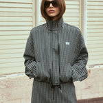 A woman wearing a black and white gingham mini jacket with a drawstring waist and sunglasses, standing in front of a beige paneled wall.