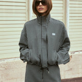 A woman wearing a black and white gingham mini jacket with a drawstring waist and sunglasses, standing in front of a beige paneled wall.