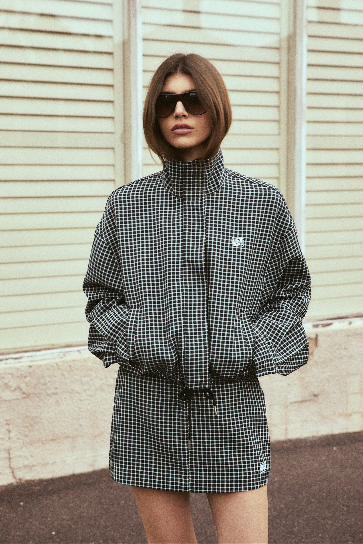 A woman wearing a black and white gingham mini jacket with a drawstring waist and sunglasses, standing in front of a beige paneled wall.
